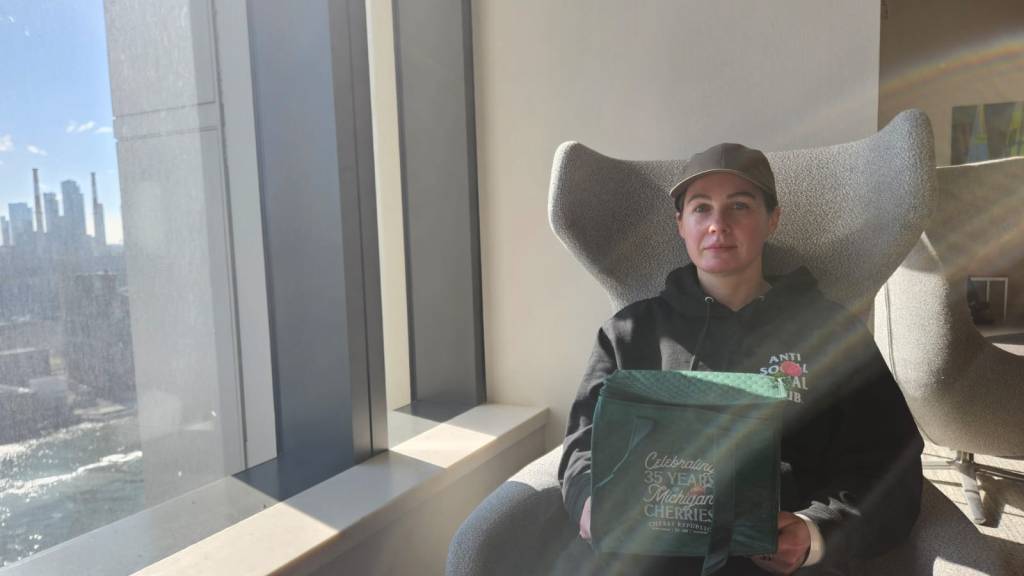 photo of me in a kind of fancy waiting room chair with the east river outside the window. i'm wearing a taupe baseball cap and an antisocial social club black hoodie with a green cooler on my lap. the cooler says celebrating 35 years of michigan cherries. i have a hospital bracelet on.